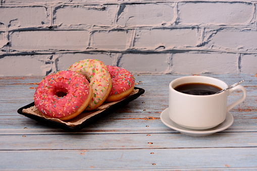 A plate with three glazed donuts with sprinkles and a cup of hot black coffee on a wooden table. Close-up.