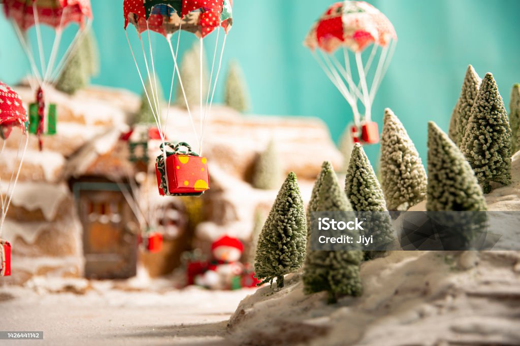 Artificial micro-model, a small fairy tale house with snow in the forest on a sunny day, with a parachute and Christmas gifts falling from the sky outside the door Sand table scenery, artificial landscape, miniature landscape, shifting axis photography. Atmospheric Mood Stock Photo