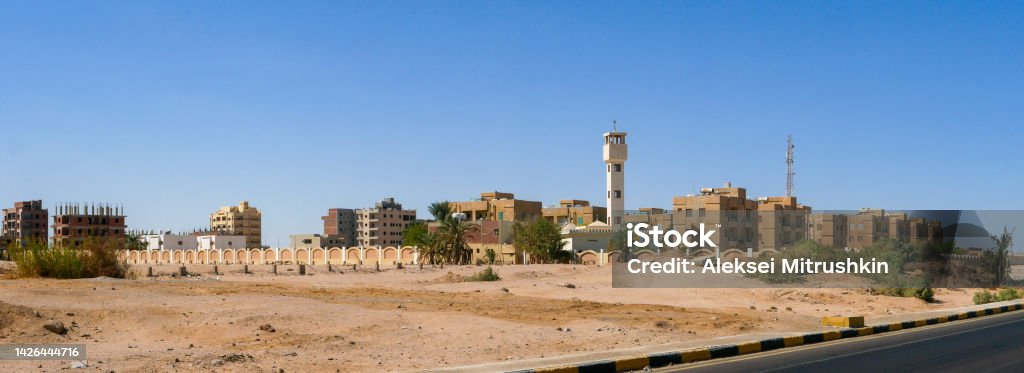 Panoramic view from the road to the low houses of local residents surrounded by desert and palm trees against the background of a blue sky. Copy space. Safaga, Egypt Panoramic view from the road to the low houses of local residents surrounded by desert and palm trees against the background of a blue sky. Copy space. Safaga, Egypt. Africa Stock Photo