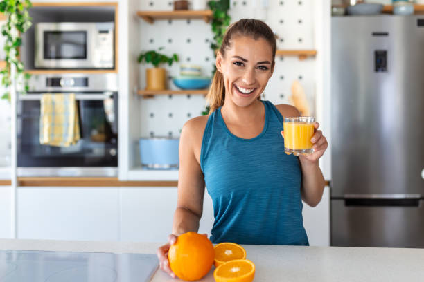 hermosa joven bebiendo jugo de naranja fresco en la cocina. dieta saludable. joven feliz con vaso de jugo y naranja en la mesa de la cocina. - drinking straw drinking juice women fotografías e imágenes de stock