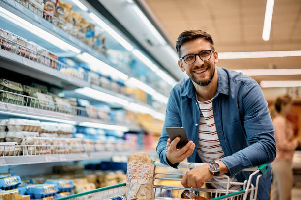 glücklicher mann, der die handy-app benutzt, während er lebensmittel im supermarkt kauft und in die kamera schaut. - markt stock-fotos und bilder