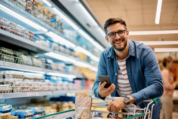 homem feliz usando aplicativo de celular enquanto compra mantimentos em supermercado e olhando para a câmera. - mercearia - fotografias e filmes do acervo