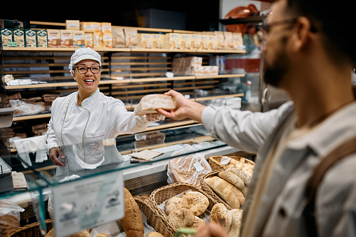 Happy baker giving fresh bread to a customer at supermarket bakery.