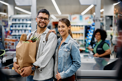 Happy couple carrying groceries in paper bag after buying in supermarket and looking at camera.