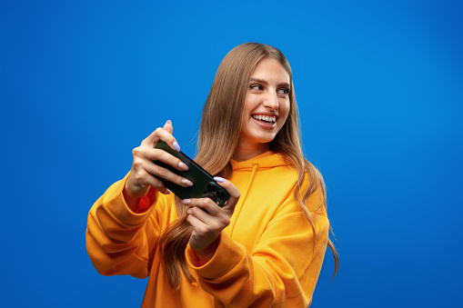 Excited young woman playing mobile video game on smartphone against blue background in studio