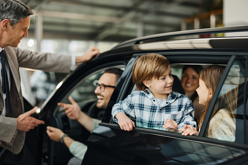Young happy family enjoying while buying a new car in a showroom. Father is talking to a salesman.