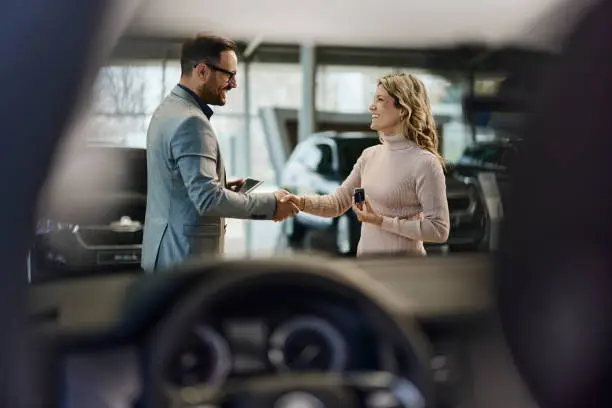 Happy salesman congratulating his female customer for buying a new car in a showroom. The view is through glass.