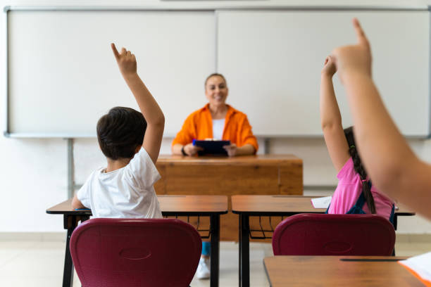 photo d’un groupe diversifié d’enfants assis dans leur classe d’école et levant la main pour répondre à une question - reading and writing child whiteboard preschool photos et images de collection