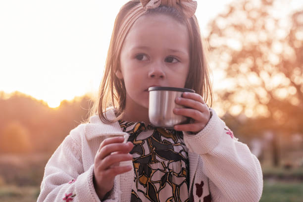 menina de 3 a 4 anos senta-se em parque fazendo piquenique em raios de pôr do sol e segura tampa térmica com chá quente na mão - preschool 2 3 years preschooler little girls - fotografias e filmes do acervo