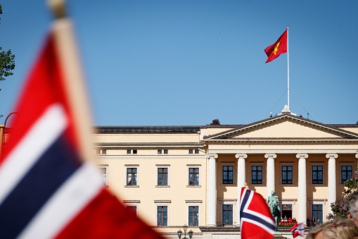 A Picture of the view towards the Norwegian Royal Palace and the Royal family standing on the Royal Balcony, waving to the crowd. The flag on the top of the roof is the Royal Standard of Norway, which is the flag of the Norwegian King, consisting of a lion holding n axe on a red field.