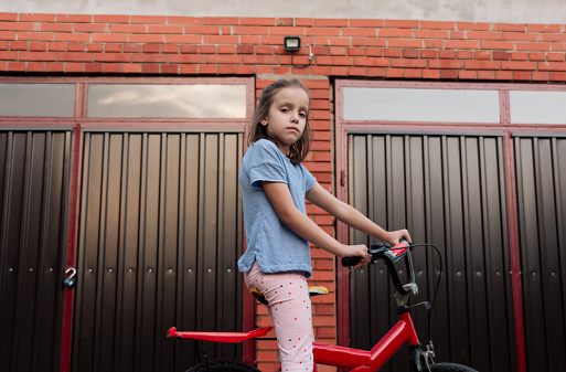 Low angle view portrait of a girl whit bike standing in the yard and looking at camera