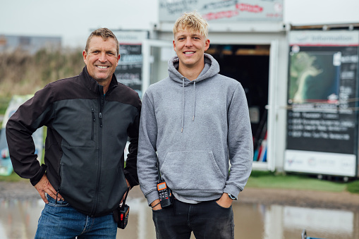 A portrait shot of a father and son standing outside a storage unit they work in together for their small water sports training business in Beadnell bay, in the North East of England.
