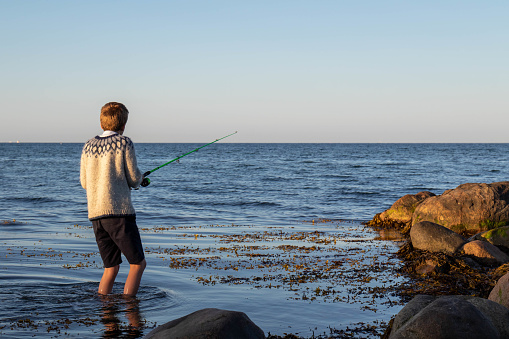 A boy wading into the water with a fishing rod. Stones on the shore, sea kelp in the water. The horizon of the ocean in the background