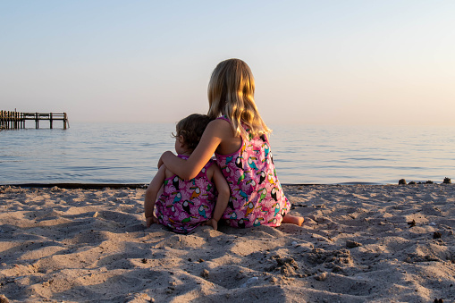 Two girls sitting on a sandy beach, looking at the calm ocean. Sisters wearing matching dresses, the older sister embracing the younger one. The horizon of the ocean and a pier in the background.