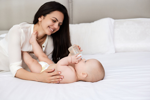 happy mother feeding her newborn baby on the bed at home. mom holding a bottle of water