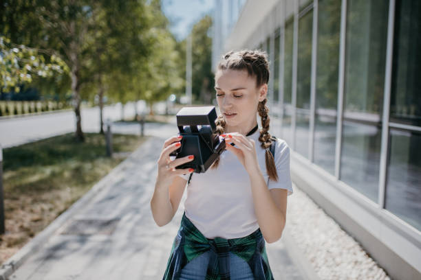 Beautiful young female student taking photos with vintage polaroid camera in front of university building