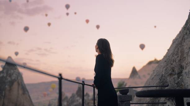 Female tourist enjoying watching hot air balloons flying in the sky at rooftop of hotel where she is staying during her vacation stock photo