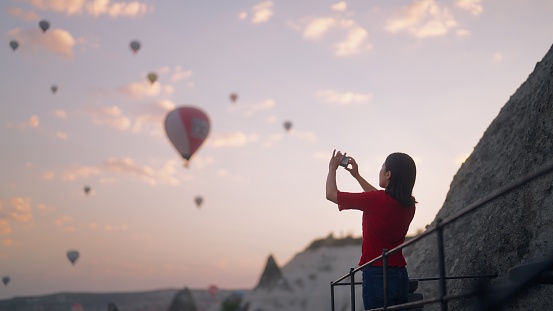 A young female tourist is taking photos of hot air balloons from the rooftop of the hotel where she is staying during her travel.