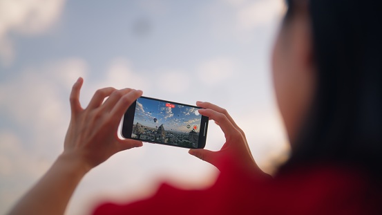 A young female tourist is taking photos of hot air balloons from the rooftop of the hotel where she is staying during her travel.