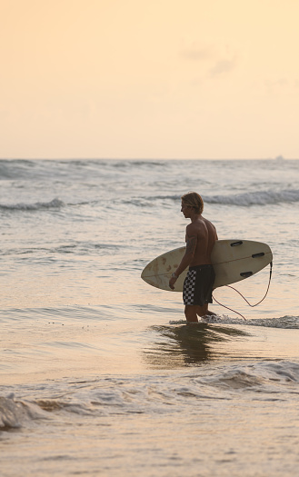 Ahangama, Sri Lanka - 04 01 2022: Lonely surfer carrying a surfboard and walking into the ocean in the evening.