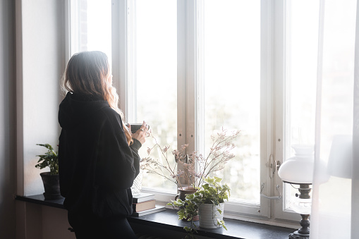 Woman standing in the window with a cup of tea in her hand. She is looking at the view through the window, taking a break from it all.