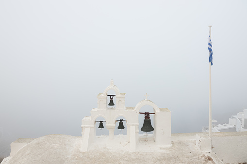 Minimalist scene of traditional Greek Orthodox church bells and Greek flag on a moody, atmospheric morning of fog and mist at the coastal village old town of Oia, Santorini, Greece.