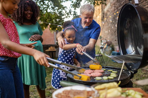 A Caucasian grandfather helping his multiracial granddaughter grill food at a back yard barbecue party