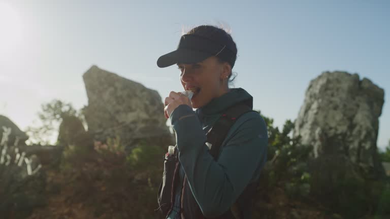 Hiker enjoying her snack on the mountain trail. Young female jogger eating an energy bar on a windy mountain path, exploring nature. Keeping energy up with healthy nutrition during endurance training.