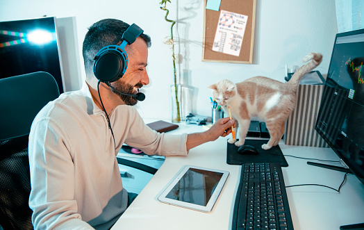 Smiling Man Customer representative with headphones working from home with his cat walking on a desk