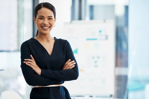 mujer de negocios segura, feliz y sonriente de pie con los brazos cruzados mientras está en una oficina con una mentalidad positiva y un buen liderazgo. retrato de un emprendedor sintiéndose motivado y orgulloso - bien vestido fotografías e imágenes de stock