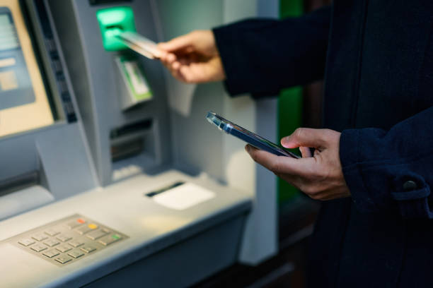 Close up of man using smart phone while withdrawing cash at ATM. stock photo