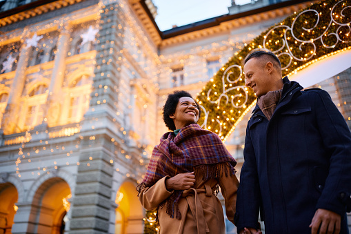 Low angle view of happy couple walking through Christmas decorated city during winter holidays. Copy space.