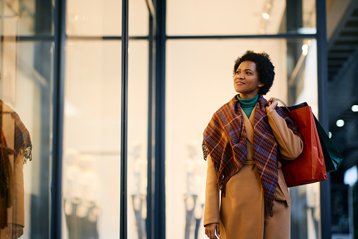 Smiling African American woman with shopping bags looking at store windows while walking through the city. Copy space.