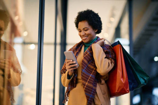 happy black woman texting on cell phone while shopping in the city. - mercadoria imagens e fotografias de stock