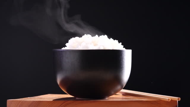 Japanese rice, Cooked rice. Close up natural steaming cooked Japanese white rice in black bowl with chopstick on black background, soft focus. Healthy Food Concept.