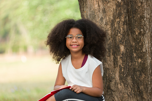 Happy African American child girl wearing glasses reading a book outdoors in the park. Kid girl learning outside at the school