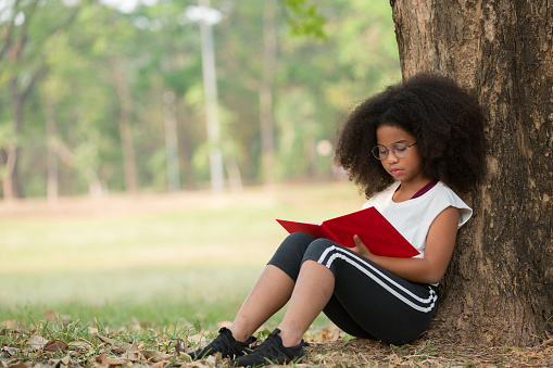 Happy African American child girl wearing glasses reading a book outdoors in the park. Kid girl learning outside at the school