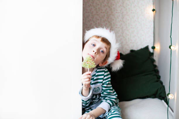 little boy wearing striped pajamas and santa claus hat sitting in a white wardrobe with a lollipop. - christmas child little boys peeking imagens e fotografias de stock