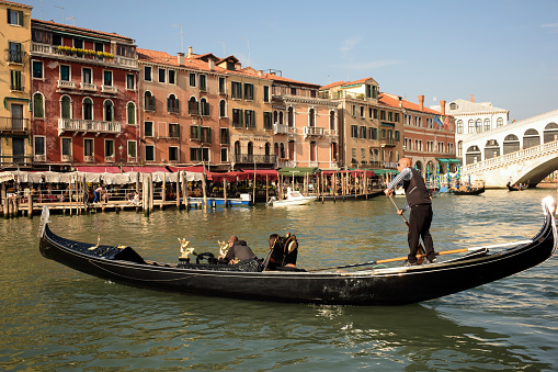 October 16, 2019, Venice, Italy: On a boat, a gondolier transports a tourist along the Grand Canal, with a building and vacationers in the background