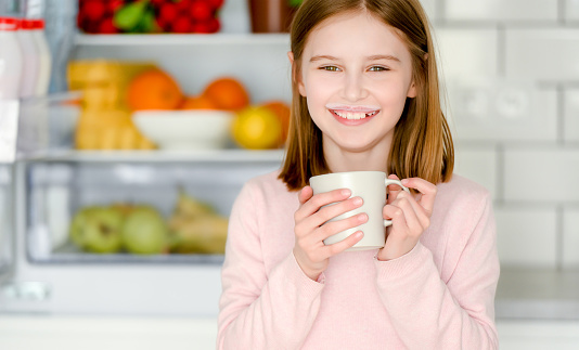 Pretty preteen girl drinking yogurt from cup mug at kitchen and smiling with milk trace over her lips. Beautiful female kid with desert at home
