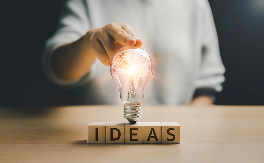 Woman's hand touching a light bulb glowing on a wooden block with Word Ideas. Light bulb. Human brain icon inside. the new concept that is innovative and inspiring Technological innovations in science and communication concepts.