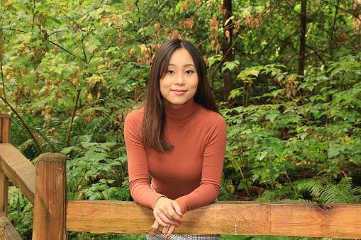 A smiling Chinese woman leaning on a wood rail fence in a public park. She is wearing a brown turtleneck sweater.