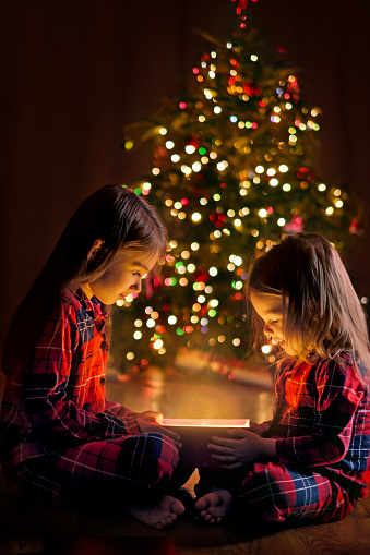 Two cute beautiful girls in checkered pajamas on a magical Christmas evening open Santa Claus' gift sitting on the floor by the Christmas tree with lights of garlands. A night of magic and miracles