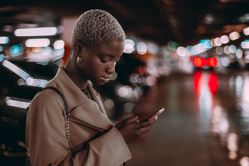 A low-key portrait with a shallow depth of field and a selective focus on a charming young African woman using her smartphone while standing in an underground parking; a copy space place on the right