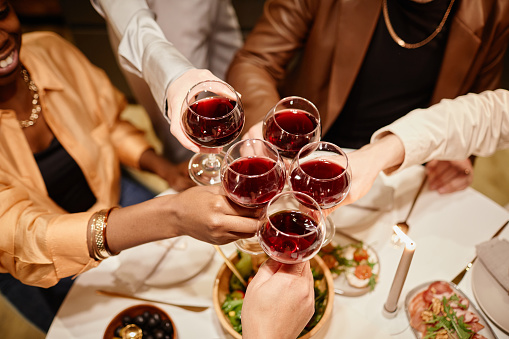 Above view of hands of young friends clinking with wineglasses over dinner table served with homemade food during celebration