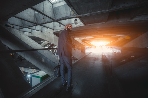 A wide-angle shot of an elegant black bald bearded businessman in a formal suit, walking through a modern futuristic covered pedestrian passageway made of concrete constructions; light at the end of the tunnel