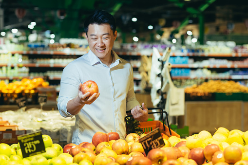 young asian man chooses and picks in eco bag apple fruit or vegetables in the supermarket. male customer standing a grocery store near the counter buys and throws in a reusable package in market