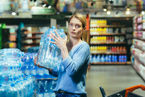 Portrait of scared woman shopper in supermarket buying water in plastic bottles and looking scared at camera