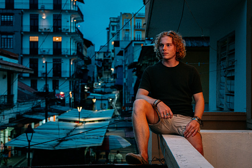 Portrait of a Comfortable, Fashionable, Handsome Young Caucasian Man Sitting on the Edge of a Balcony Overlooking the Night Life Streets of Panama City at Dusk in the Summer