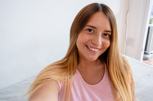 beautiful young caucasian venezuelan woman smiling takes selfie indoors in her room, sitting on her bed looking at the happy vamara dressed in pink with dyed blonde hair and black eyes.